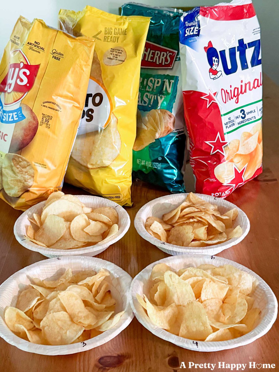 Bag of classic potato chips by Lays, Great Value, Herrs, and Utz lined up in front of numbered paper bowls filled with chips for a potato chip taste test