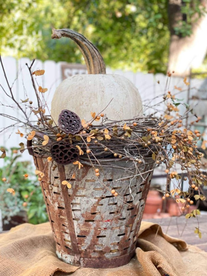 white pumpkin sitting on some dried branches and vines inside a rustic olive bucket for a fall centerpiece