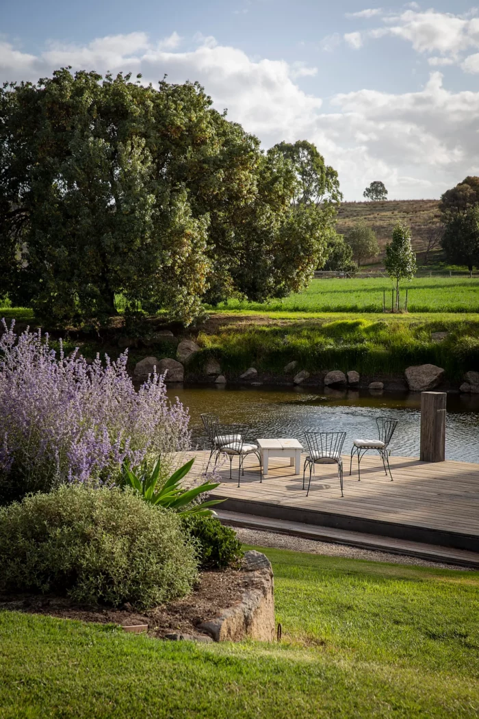 metal chairs and coffee table sitting on a wood dock next to a creek in Australia with green trees and grass on either side of the creek in a delightfully serene scene