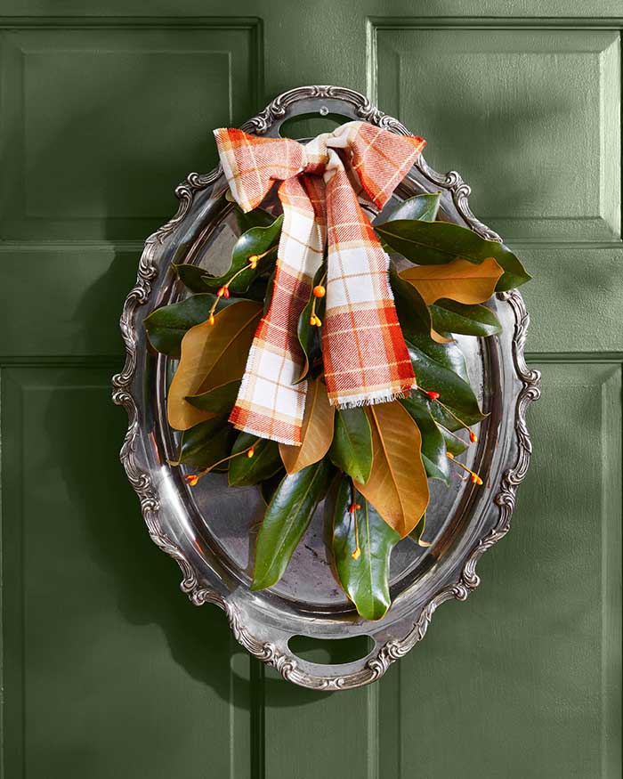 fall wreath made from an oval-shaped silver platter with magnolia leaves and an orange plaid ribbon. the wreath is hanging on a green door.